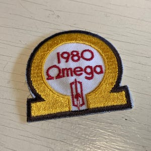 Peut inclure: Écusson brodé avec une bordure dorée et un centre blanc. L'écusson porte l'inscription "1980 Omega" en rouge, avec un emblème stylisé en dessous. L'écusson a une bordure marron et une extension rectangulaire.