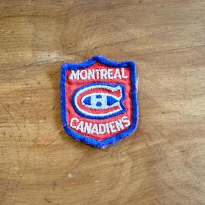 May include: A shield-shaped embroidered patch featuring a red background with a blue border. The text MONTREAL CANADIENS is stitched in white, surrounding a central blue and white C logo with an H inside. The patch has a visible loose red thread.