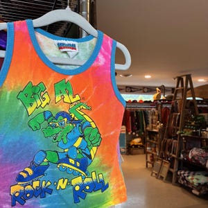 Könnte beinhalten: Ein farbenfrohes Tanktop mit Batik-Effekt, das ein Cartoon-Alligator auf Rollschuhen zeigt. Der Text "BIG AL" und "ROCK N' ROLL" sind deutlich zu sehen. Das Oberteil hat einen blauen Rand und hängt an einem weißen Kleiderbügel.
