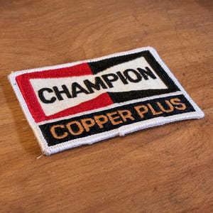 May include: Rectangular embroidered patch featuring the word "CHAMPION" in black on a white background, with red and black sections. The text "COPPER PLUS" is below. The patch has a white border.