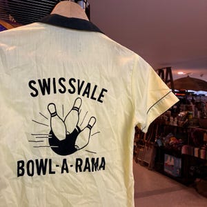 May include: A light yellow bowling shirt with a black collar and trim. The back of the shirt features the text "SWISSVALE BOWL-A-RAMA" above a graphic of bowling pins and a bowling ball.