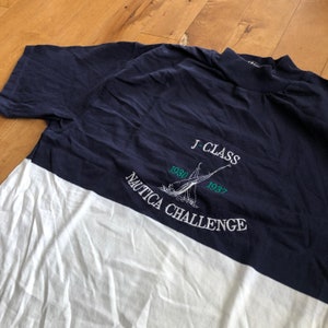 Könnte beinhalten: Ein marineblaues und weißes T-Shirt mit einem Segelboot-Motiv und dem Text "J-Class Nautica Challenge 1930 1937".