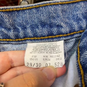 May include: Close-up of the inside of blue denim jeans. The image shows a white tag with text and numbers, including "29/30". The jeans have yellow stitching and are slightly faded.