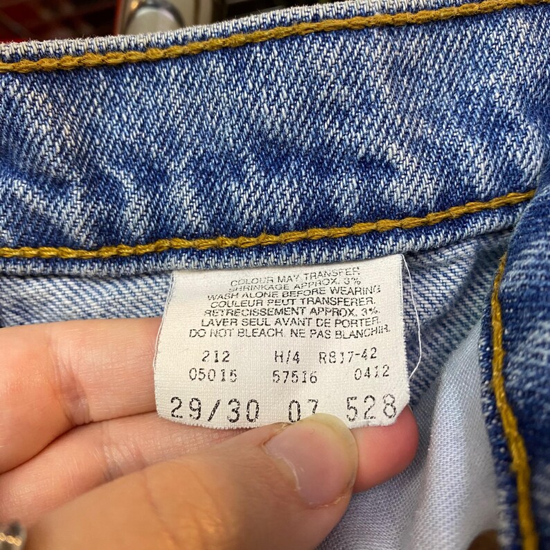 May include: Close-up of the inside of blue denim jeans. The image shows a white tag with text and numbers, including "29/30". The jeans have yellow stitching and are slightly faded.