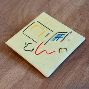 Peut inclure: Sous-verre carré jaune clair avec un graphisme d'ordinateur rétro en noir, rouge, jaune et bleu. Le design comprend un moniteur d'ordinateur stylisé, un clavier et le logo Apple. Le dessous de verre a une finition brillante.