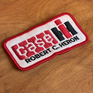 May include: Embroidered patch with a white background and red border. The patch features the word "case" in red, stylized text, and the letters "IH" in red and black. The text "ROBERT C. HERON" is below the logo.
