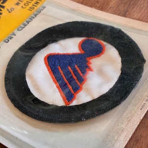 May include: A circular patch with a black border, white centre, and a blue and orange emblem. The patch is in a clear plastic package. The emblem appears to be a stylized figure or logo. The patch is likely for clothing or accessories.