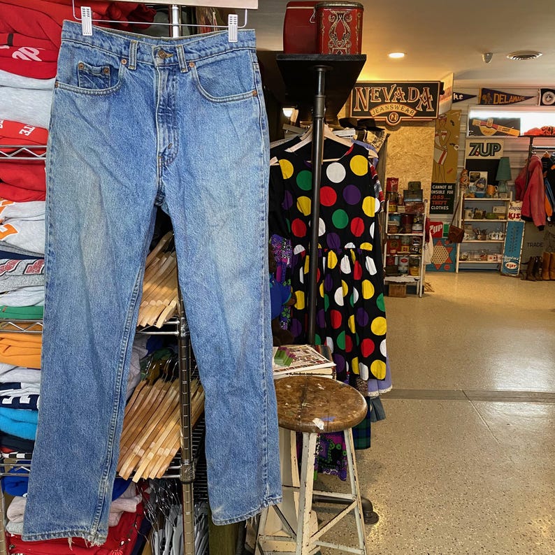 May include: Light blue denim jeans hanging on a metal rack. The jeans have a classic five-pocket design and a straight leg fit. The jeans are displayed in a vintage clothing store.
