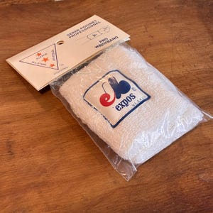 May include: A white terry cloth wristband, packaged in clear plastic, with a label featuring a red cherry and blue design. The packaging includes text in French and English, indicating it's a professional wristband.