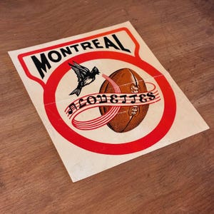 May include: A vintage Montreal Alouettes sticker featuring a football, a bird, and the team name. The sticker has a red and white border with the word "MONTREAL" at the top. The football is brown, and the bird is black.