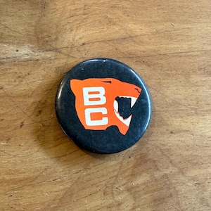 May include: A circular pinback button featuring a black background with a stylised orange graphic of a roaring panther head. The letters BC are printed in white inside the panther graphic, which has an open mouth showing sharp teeth.