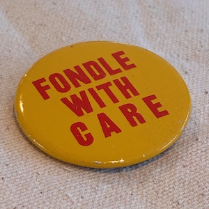 May include: A circular yellow badge with the red text "FONDLE WITH CARE". The badge has a slightly distressed look. The background is a textured, off-white fabric, possibly linen or cotton. The badge is a vintage item.