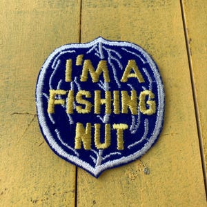 Op de afbeelding: Een marineblauwe patch met de tekst "I'M A FISHING NUT" in gele geborduurde letters. De patch heeft een witte rand en een bladachtig ontwerp rond de rand.
