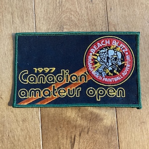 May include: A black and green embroidered patch with the text "1997 Canadian amateur open" and a red circle with the text "Beach Blitz" and a graphic of a person in a paintball suit.