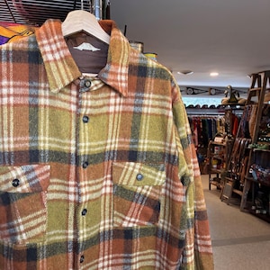 May include: A vintage plaid shirt in shades of orange, yellow, and brown, with black buttons and two front pockets. The shirt is hanging on a wooden hanger, showcasing its classic design and textured fabric.