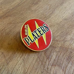 May include: A circular red enamel pin featuring the word "PLATERS" in black with a gold outline. The word "GUELPH" is arched above the design. Two yellow lightning bolts are behind the word "PLATERS". The pin has a gold border.