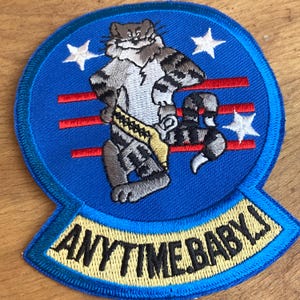 May include: Embroidered patch with a raccoon character in a blue circle, red stripes, and white stars. The raccoon wears a yellow belt and has a lightning bolt on its chest. The text "ANYTIME BABY!" is displayed in gold lettering.