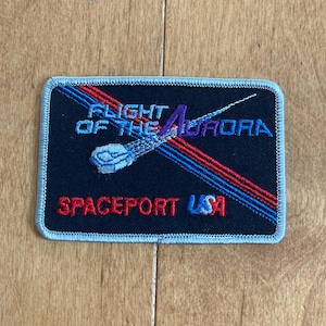 Peut inclure: Un écusson brodé noir avec des coutures bleues et rouges. L'écusson représente un vaisseau spatial stylisé avec le texte "Flight of the Aurora" et "Spaceport USA".