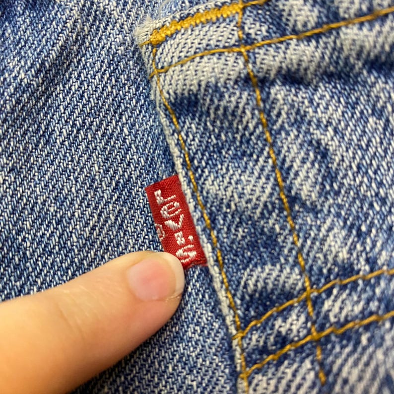May include: Close-up of blue denim fabric with yellow stitching. A small red tag with the word "Levi's" in white is visible. The tag is attached to the denim.