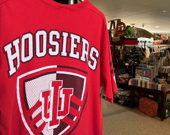 Könnte beinhalten: Ein rotes T-Shirt mit dem Wort "HOOSIERS" in weißen Blockbuchstaben. Darunter befindet sich eine schildförmige Grafik mit den Buchstaben "IU" in Weiß und Rot. Das T-Shirt hängt an einem Holzkleiderbügel.