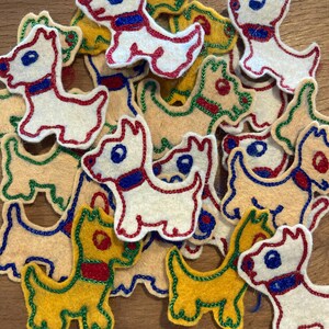 Peut inclure: Une collection d'appliqués en forme de chien en feutre de différentes couleurs. Les chiens sont principalement blancs, jaunes et beiges, avec des détails brodés rouges, bleus et verts. Les appliqués sont dispersés sur une surface en bois.