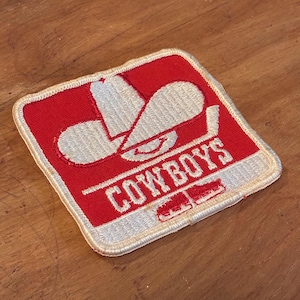 May include: A square, embroidered patch with a red background and a white cowboy hat design. The word "COWBOYS" is written in white block letters. The patch has a white border.