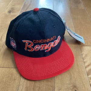 Peut inclure: Une casquette de baseball noire et rouge avec les mots "Cincinnati Bengals" brodés en orange sur le devant. La casquette a une visière noire et une couronne rouge. Le logo de la NFL est visible sur le côté de la casquette.