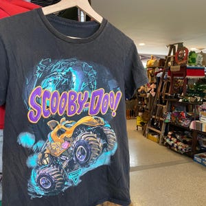 May include: Black t-shirt featuring the text "Scooby-Doo" in purple and yellow. The graphic includes a monster truck with the words "Scooby Doo" on the side, set against a blue and green background.