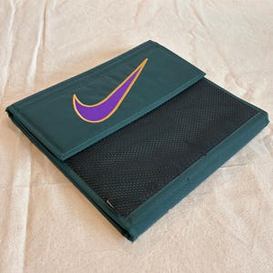 May include: Dark green document holder featuring a purple and gold swoosh logo. The holder has a black mesh pocket and a flap closure. The item is made of a durable material and is designed to protect documents.