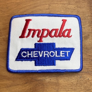 May include: A rectangular patch with a white background, featuring the red word "Impala" above a blue Chevrolet logo. The patch is bordered with blue stitching, showcasing a vintage automotive emblem.