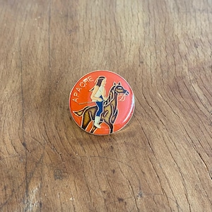 Könnte beinhalten: Eine runde, orangefarbene Emaille-Anstecknadel mit goldfarbenem Rand. Die Anstecknadel zeigt ein Cartoon-Bild einer Person, die auf einem Pferd reitet. Der Text "APACHE" ist um den Rand der Anstecknadel herum geschrieben.