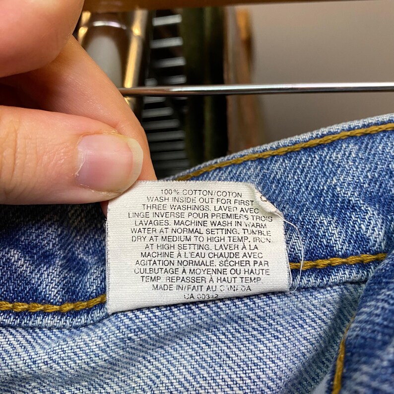May include: Close-up of a white tag with care instructions attached to blue denim jeans. The tag includes text in English and French, detailing washing and drying instructions. The jeans have yellow stitching.