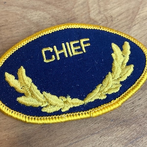 Op de afbeelding: Een marineblauwe patch met het woord "CHIEF" geborduurd in goud. De patch heeft een gouden lauwerkransontwerp en een gouden rand. De patch is ovaal van vorm.