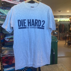 May include: A gray t-shirt with the text "DIE HARD 2" in black lettering and "DIE HARDER" in smaller black lettering below. The shirt is on a hanger.