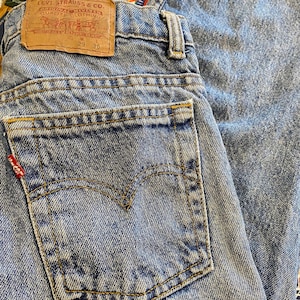 May include: Close-up of light blue denim jeans with a classic five-pocket design. The jeans feature a brown leather patch with the Levi Strauss & Co. logo and size details. The back pocket has a red tag. The waist size is 29 inches, and the length is 30 inches.