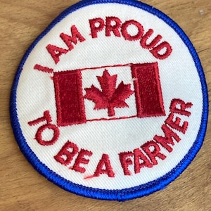 Op de afbeelding: Geborduurde patch met een witte achtergrond, blauwe rand en de tekst "I AM PROUD TO BE A FARMER" in het rood. De patch heeft in het midden een rood-wit Canadees vlagontwerp.