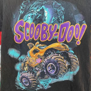 May include: Black t-shirt featuring a graphic of the Scooby-Doo monster truck. The design includes the Scooby-Doo logo in purple and yellow, a yellow and orange monster truck, and the Monster Jam logo.