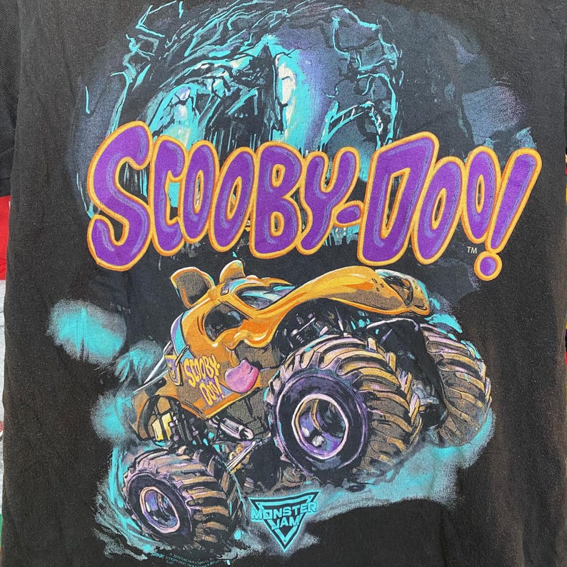 May include: Black t-shirt featuring a graphic of the Scooby-Doo monster truck. The design includes the Scooby-Doo logo in purple and yellow, a yellow and orange monster truck, and the Monster Jam logo.