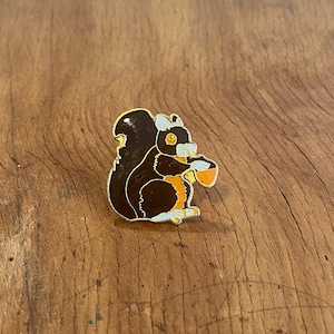 May include: An enamel pin featuring a black and orange squirrel holding an acorn. The squirrel is sitting on its haunches and has a white belly.