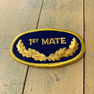 May include: An oval navy blue embroidered patch with the text "1ST MATE" in gold. The patch has a gold border and gold laurel leaves. The patch is suitable for sewing onto clothing or bags.