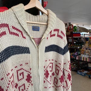 May include: A cream-coloured knitted cardigan with a zip closure. The cardigan features a geometric pattern in red and black. The cardigan has a collar and long sleeves.