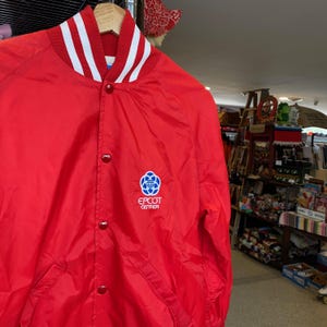 May include: Red satin bomber jacket with white stripes on the collar and a blue and white embroidered logo that says "EPCOT CENTER".