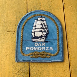 Op de afbeelding: Een blauwe geborduurde patch met een zeilschip met witte zeilen en donkerblauwe details. De tekst "DAR POMORZA" is wit geborduurd. De patch is omlijst door een gouden touwontwerp.