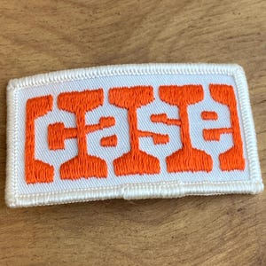 May include: A rectangular white patch with an orange embroidered design. The design features the word "CASE" in a stylized, bold font. The patch has a white border and is likely an emblem or logo.