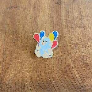 Op de afbeelding: Een blauwe beer met een feestmuts houdt drie ballonnen vast, een rode, een blauwe en een gele.