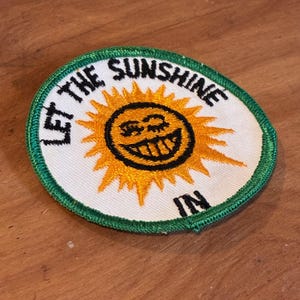 Op de afbeelding: Een ronde patch met een lachend zonontwerp. De zon is geel met een zwarte omtrek en een lachend gezicht. De tekst "LET THE SUNSHINE IN" is in het zwart gedrukt. De patch heeft een witte achtergrond en een groene rand.