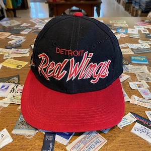 May include: A black and red baseball cap with the text "DETROIT Red Wings" in white and red script. The cap has a red brim and a red button on top. The hat is sitting on a wooden surface.