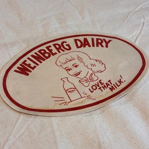 May include: Oval patch with red trim and text "WEINBERG DAIRY" in red. A vintage illustration of a woman with a bow and a milk bottle is in the center. The text "LOVE THAT MILK!" is also in red.
