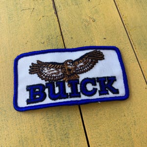 Könnte beinhalten: Gestickter Aufnäher mit einem braunen und weißen Adler mit ausgebreiteten Flügeln, über dem blau gestickten Wort "BUICK". Der Aufnäher hat einen weißen Hintergrund und einen blauen Rand. Der Aufnäher ist rechteckig.