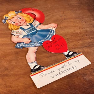May include: A vintage Valentine's Day card depicting a cartoon girl in a blue dress and black shoes. The card includes a red heart with the text "Stop Stringing my heart!" and a base with the text "Promise you'll be my VALENTINE!"